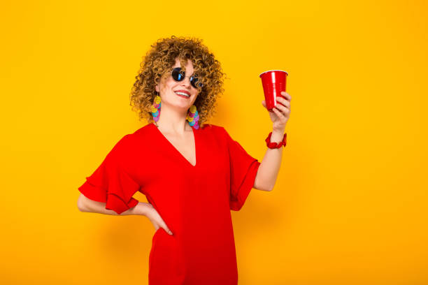 Attractive woman with short curly hair and drink Portrait of a white woman with afrro curly hairstyle in red dress and sunglasses holding red plastic cup with drink isolated on orange background with copyspace celebration concept. red party cup stock pictures, royalty-free photos & images