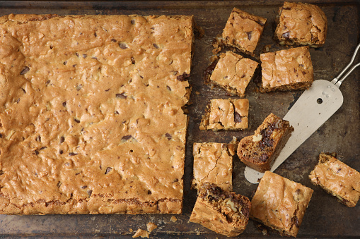 An overhead close up horizontal photograph of a freshly baked and partially sliced blondie bar cake on a baking tray.