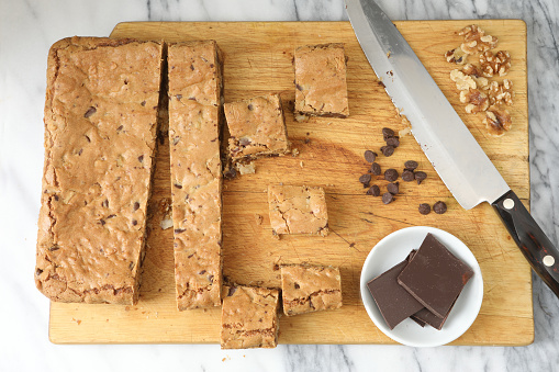 An overhead close up horizontal photograph of a partially sliced blondie bar cake and a knife on a wooden cutting board.