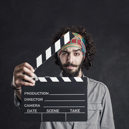 Young hipster man holding a clapperboard over black bacground, studio shot