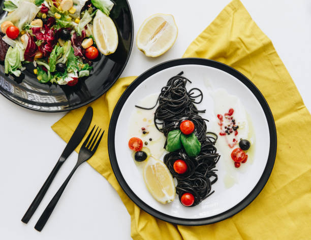 istock Pasta with squid ink and salat on white table with yellow napkin. Top view, flat lay 927736924