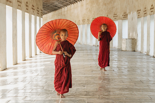 Burmese buddhist novice monks in their typical religious veils walking together along monastery archway holding their typical red parasols. Bagan, Mandalay Region, Myanmar.