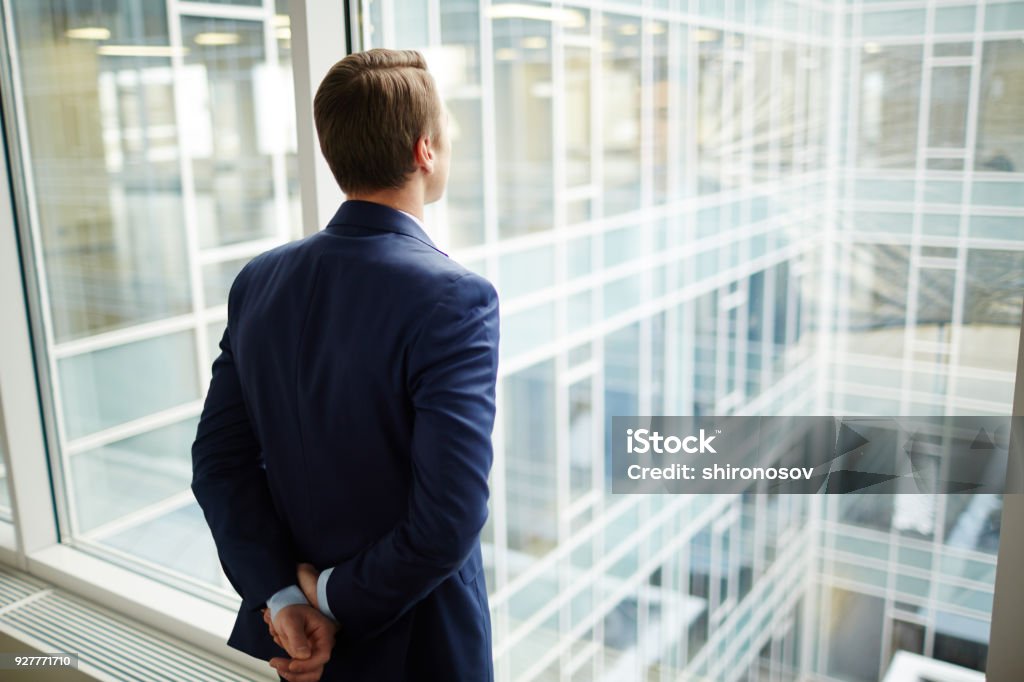 Man by window Rear view of elegant businessman standing by window of new office and looking through it Businessman Stock Photo