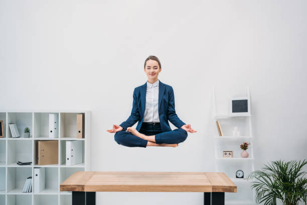 sorridente giovane imprenditrice con gli occhi chiusi meditando mentre levitare sul posto di lavoro - gambe incrociate foto e immagini stock