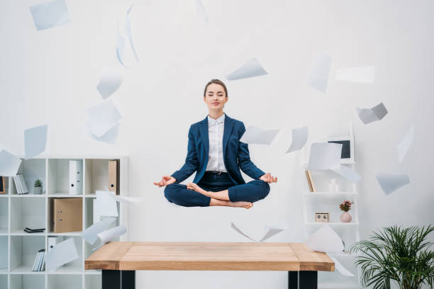 jovem empresária a sorrir com os olhos fechados, meditando enquanto levitando no local de trabalho com papéis - lotus position - fotografias e filmes do acervo