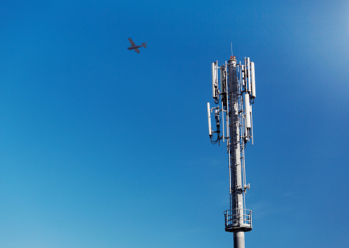 A mobile phone communications tower stands against a clear blue sky as a light aircraft flies past, high in the sky.