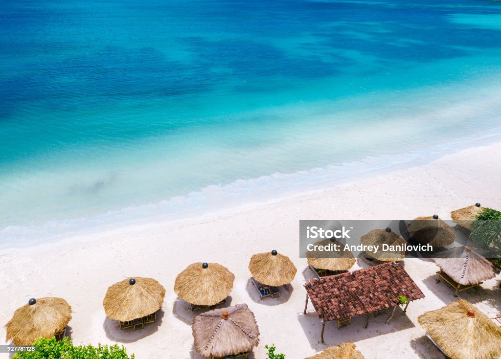 Straw beach umbrellas and blue ocean. Beach scene from above Lovely sunny day on the beach. Lombok island, drone shot Bali Stock Photo
