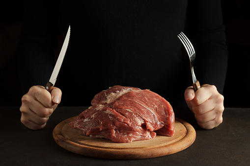 Female hands hold fork and knife and ready to eat raw meat on the wooden cutting board on dark background.