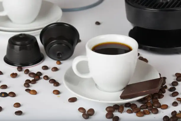 Cup of short espresso,coffee capsules, piece of black chocolate and beans on a white surface. Closeup