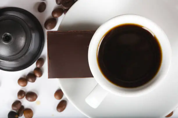 Cup of short espresso,coffee capsules, piece of black chocolate and beans on a white surface. Closeup