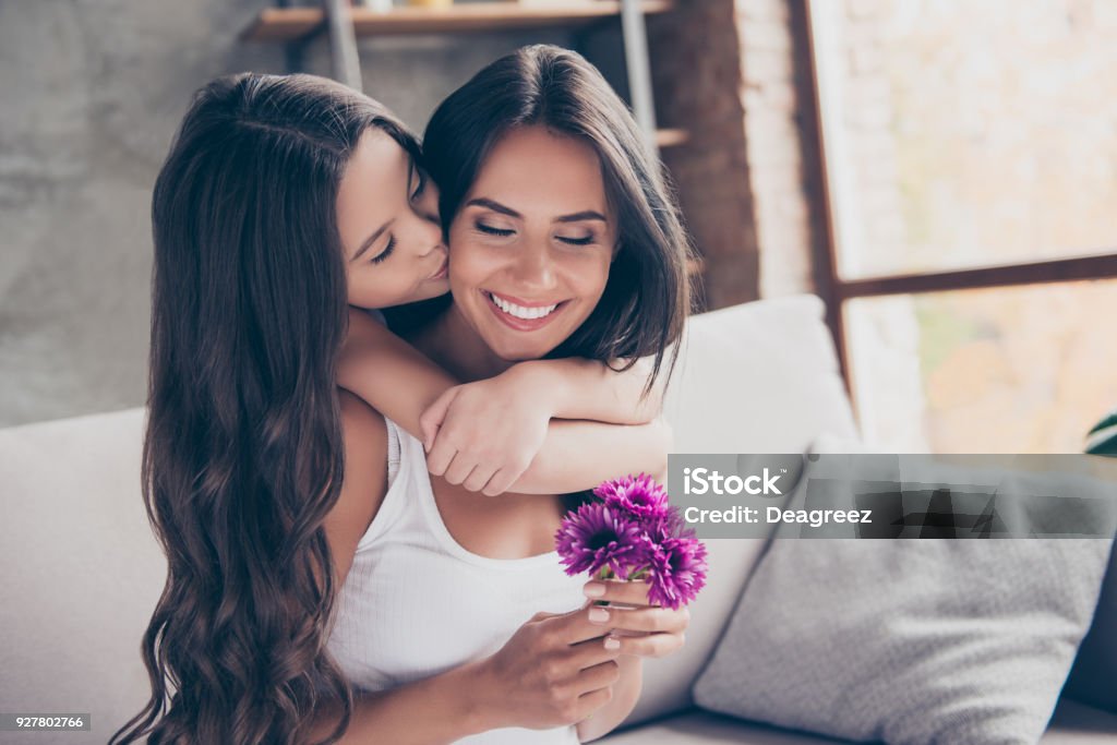 Concept of women's day! Amazed cheerful joyful charming mum is holding nice flowers and is receiving a kiss from her little cute daughter with long curly hair Mother Stock Photo