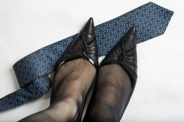 Womans feet in black transparent tights and high heels on mans tie. Gray surface