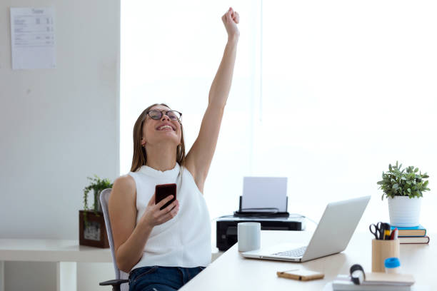 joven mujer de negocios celebrando una victoria mientras trabaja con el teléfono móvil en la oficina. - eufórico fotografías e imágenes de stock