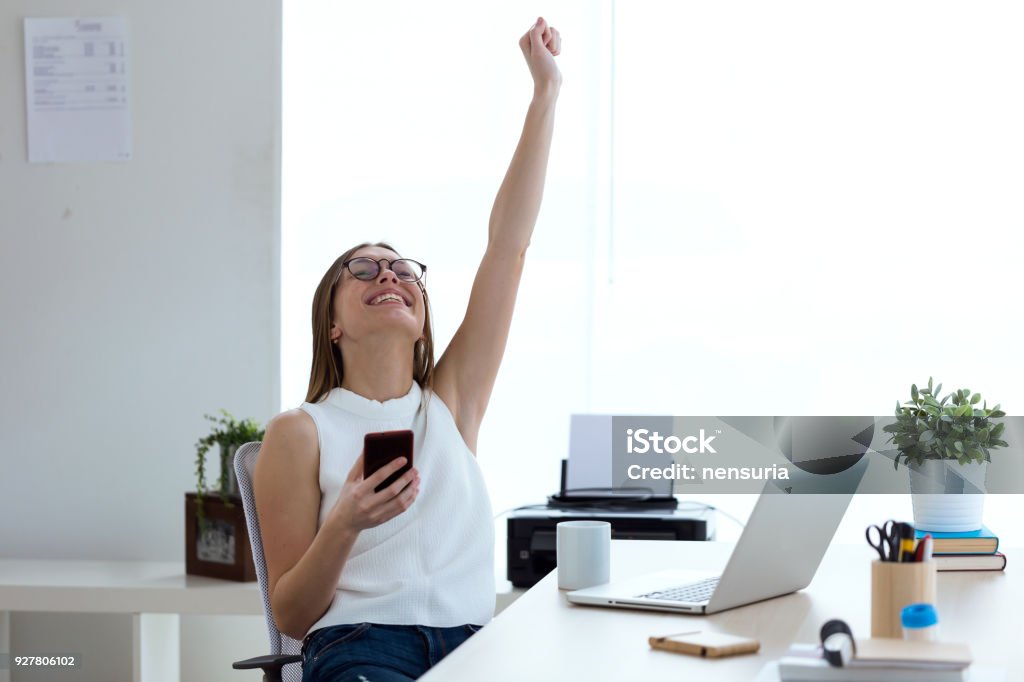 ビジネスの若い女性がオフィスで携帯電話を操作しながら、勝利を祝います。 - 成功のロイヤリティフリーストックフォト