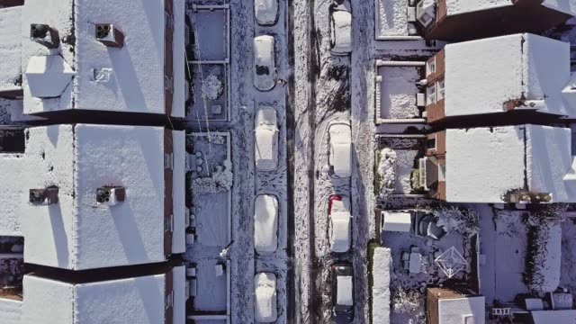 Aerial flight down a traditional UK terraced house street that’s covered in snow after a blizzard the night before.