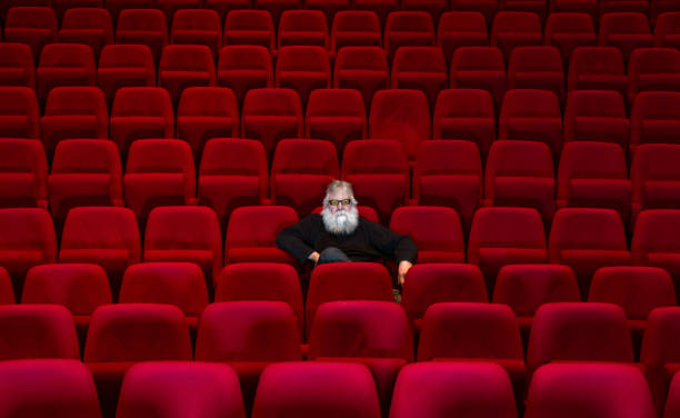 One man with white beard sits in Empty cinema or theatre with comfortable red seats