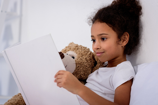 adorable african american kid reading book and hugging teddy bear at home