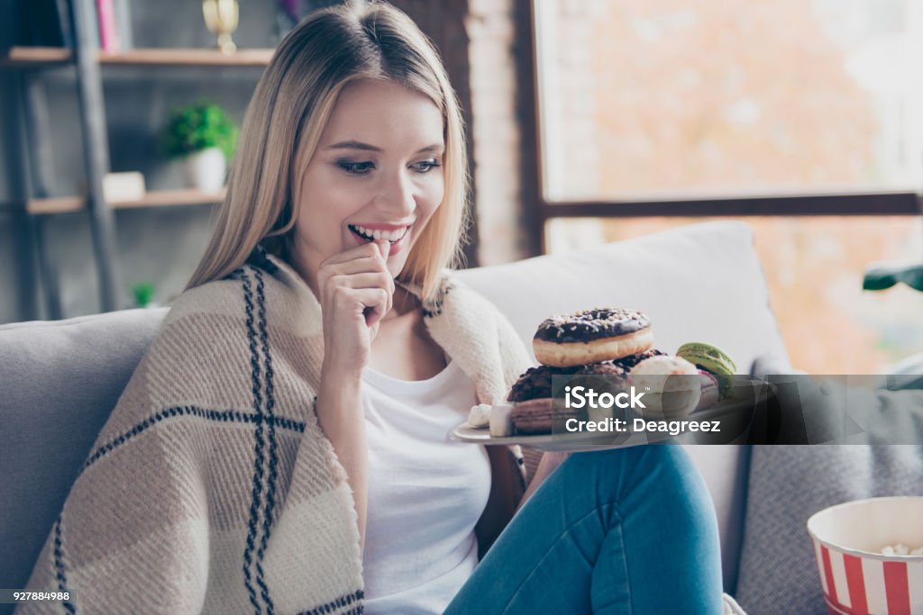 Portrait of beautiful emotional charming attractive sweet toothy woman sitting on sofa in living room, holding plate of donuts and macaroons, looking exciting satisfied Eating Stock Photo