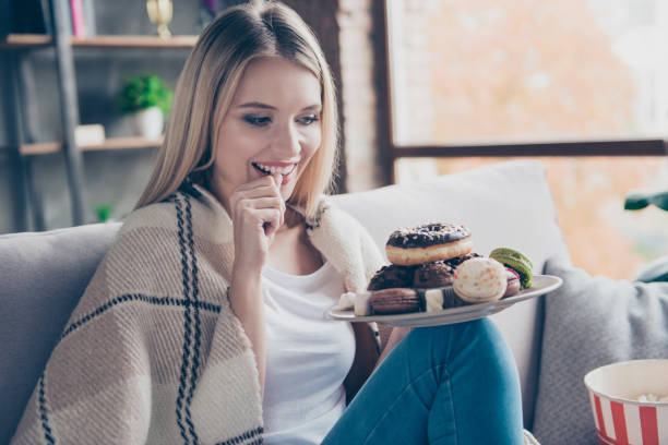 Portrait of beautiful emotional charming attractive sweet toothy woman sitting on sofa in living room, holding plate of donuts and macaroons, looking exciting satisfied Portrait of beautiful emotional charming attractive sweet toothy woman sitting on sofa in living room, holding plate of donuts and macaroons, looking exciting satisfied over eating stock pictures, royalty-free photos & images