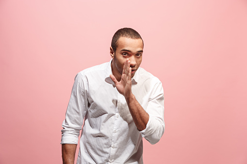 Secret, gossip concept. Young man whispering a secret behind his hand. Afro businessman isolated on trendy pink studio background. Young emotional man. Human emotions, facial expression concept.