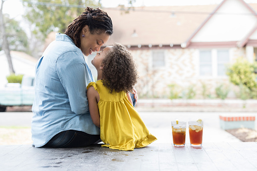 In this view from behind, a mid adult mother and her preschool age daughter sit on their front porch steps and rub noses.  There are two glasses of iced tea beside them.