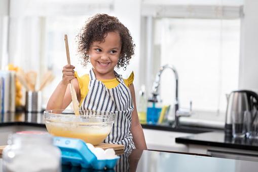 An adorable preschool age girl stands in a kitchen and mixes a bowl of batter.  She looks in the distance and smiles.