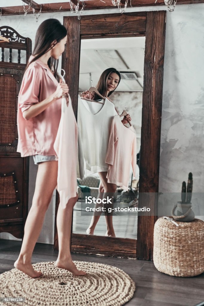 Hard choice. Full length of attractive young woman choosing the blouse and smiling while standing in front of the mirror at home Women Stock Photo