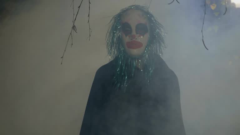 Crazy evil halloween clown coming up from a foggy wood and doing a ritual dance to frighten people