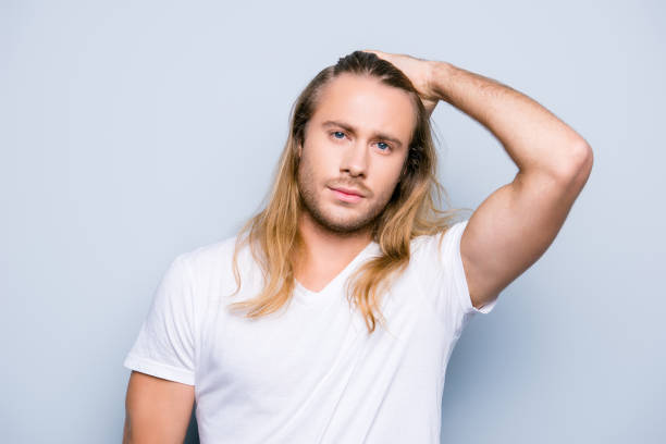 istock Handsome brutal man with bristle is combing his long blonde hair using his hand recently  he has visited a barber shop, he is isolated on grey background 927915324
