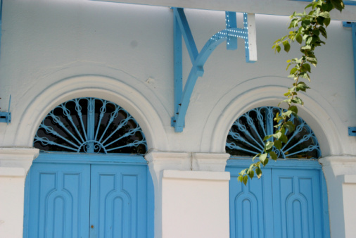 Wooden door closed and three glass windows with open shutters blue color on white wall background. Cyclades island house front view, Greek traditional architecture.