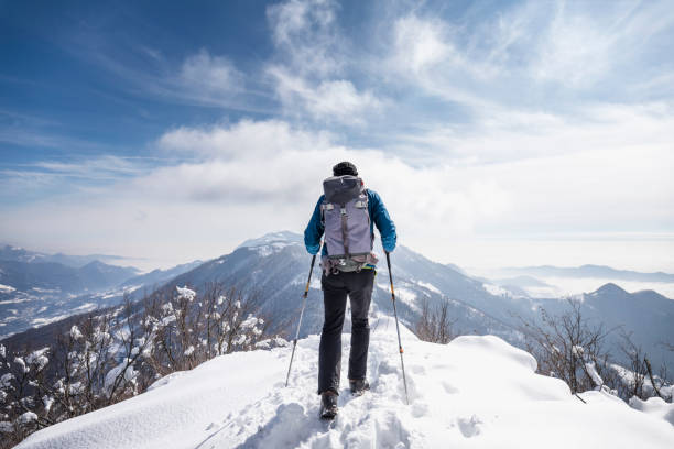 Beautiful woman dressed stylishly in vivid yellow jacket and purple pants, walking along a snowy winter trail using trekking poles in sunny day.