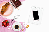 Beautiful flatlay arrangement with croissant, cup of coffee, fresh strawberries, smartphone with black copyspace and other business accessories: concept of busy morning breakfast, white background.