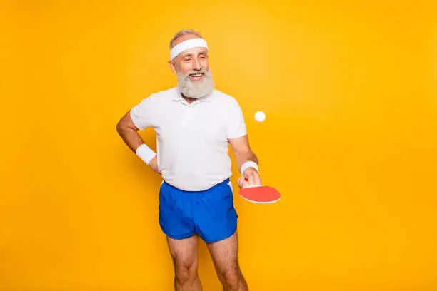Competetive emotional cool active comic grandpa with beaming grin, with table tennis equipment. Healthcare, weight loss, bodycare lifestyle, wearing blue sexy shorts, so hot!