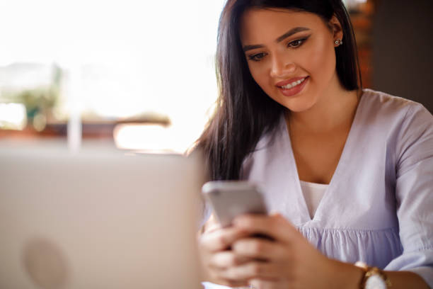 istock Smiling young woman using mobile phone and laptop at a cafe 928025434