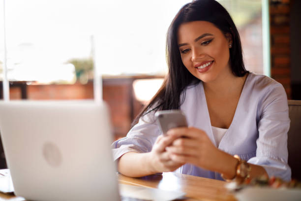 istock Smiling young woman using mobile phone and laptop at a cafe 928027644