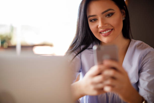 istock Smiling young woman using mobile phone and laptop at a cafe 928027650