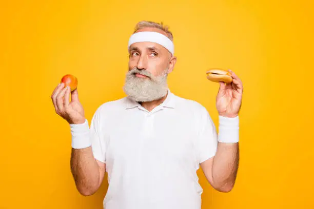 Photo of Athlete cool grandpa holds forbidden junkfood cheeseburger and fruit. Weightloss, decision, motivation, healthcare, strength, prohibition, workout, gym, regime, bodycare, calories lifestyle