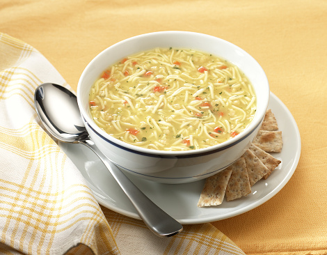 Bowl of chicken noodle soup on yellow place mat with saucer, crackers, spoon, and napkin.