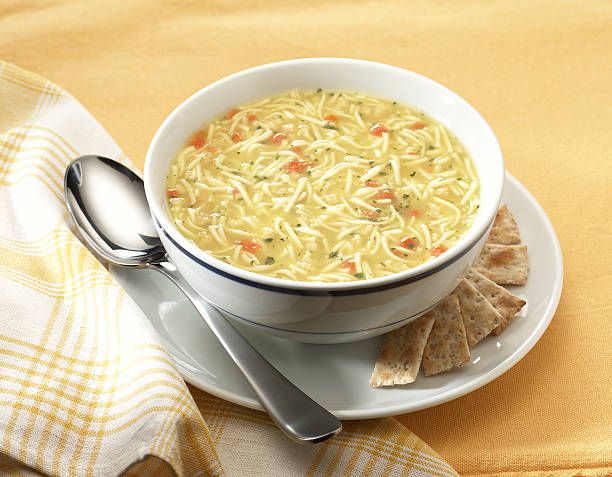 Chicken noodle soup in cup with saucer and spoon and napkin Bowl of chicken noodle soup on yellow place mat with saucer, crackers, spoon, and napkin. noodle soup photos stock pictures, royalty-free photos & images