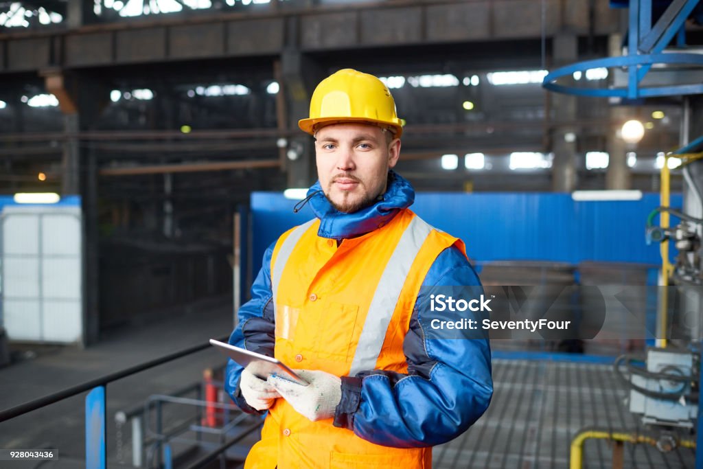 Handsome Worker Using Digital Tablet Waist-up portrait of handsome young worker wearing uniform and hardhat looking at camera while holding digital tablet in hands in order to operate machine at production department of modern plant. Adult Stock Photo