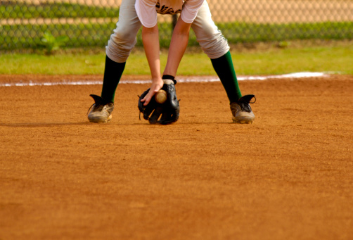 a DSLR of Baseball Player Playing in Live Baseball Game while Fielding Baseball. the baseball player is playing in a baseball uniform with baseball glove and cleats. the baseball player is playing in the infield next to third base on the baseball field. the baseball player is an athlete playing in the live sporting event. there is dirt in the infield on this baseball field. the game is being played during the day. the lighting is natural lighting of sunlight. 