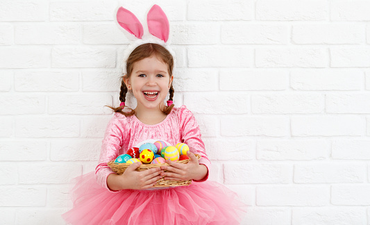 Happy easter! funny child girl with rabbit ears and a basket  of eggs at blank white wall