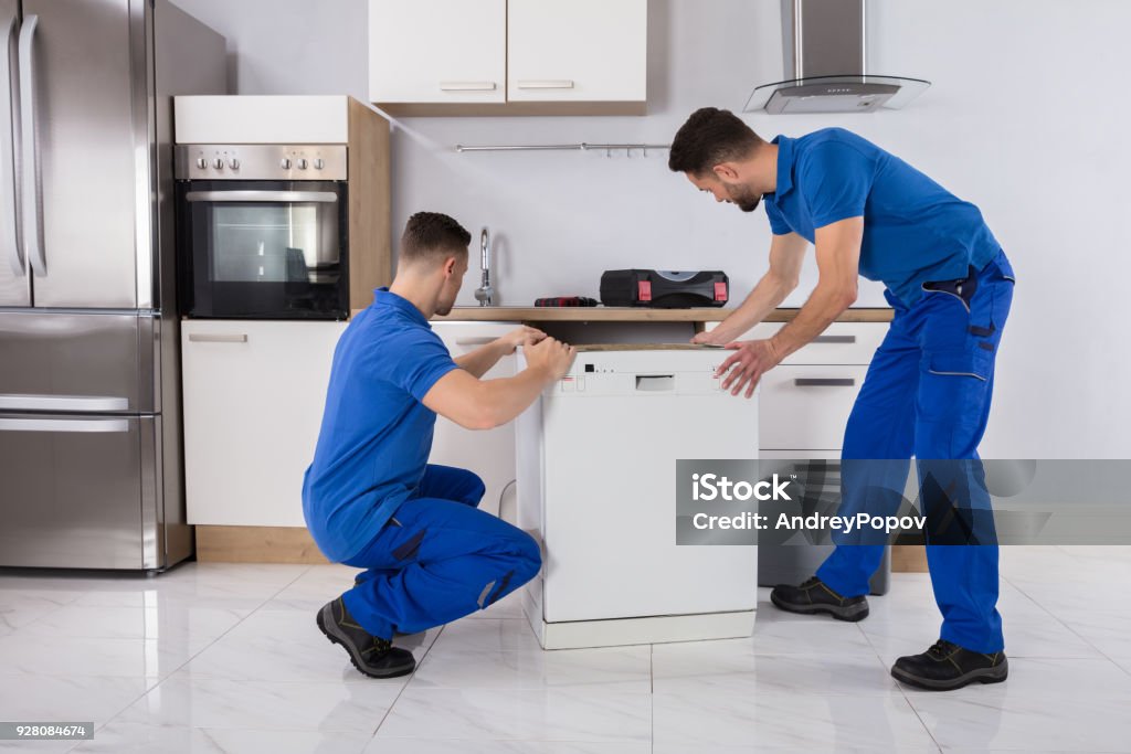 Two Movers Placing Dishwasher In Kitchen Two Young Male Movers In Uniform Placing Dishwasher In Kitchen Installing Stock Photo