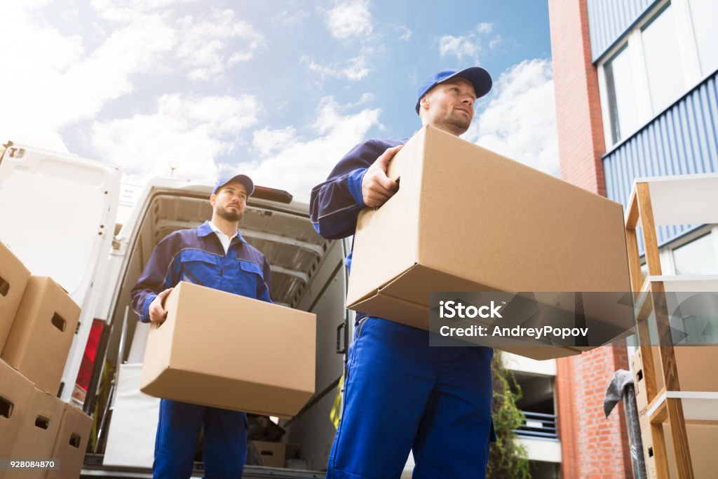 Close-up Of Two Delivery Men Carrying Cardboard Box Close-up Of Two Young Delivery Men Carrying Cardboard Box In Front Of Truck Relocation Stock Photo