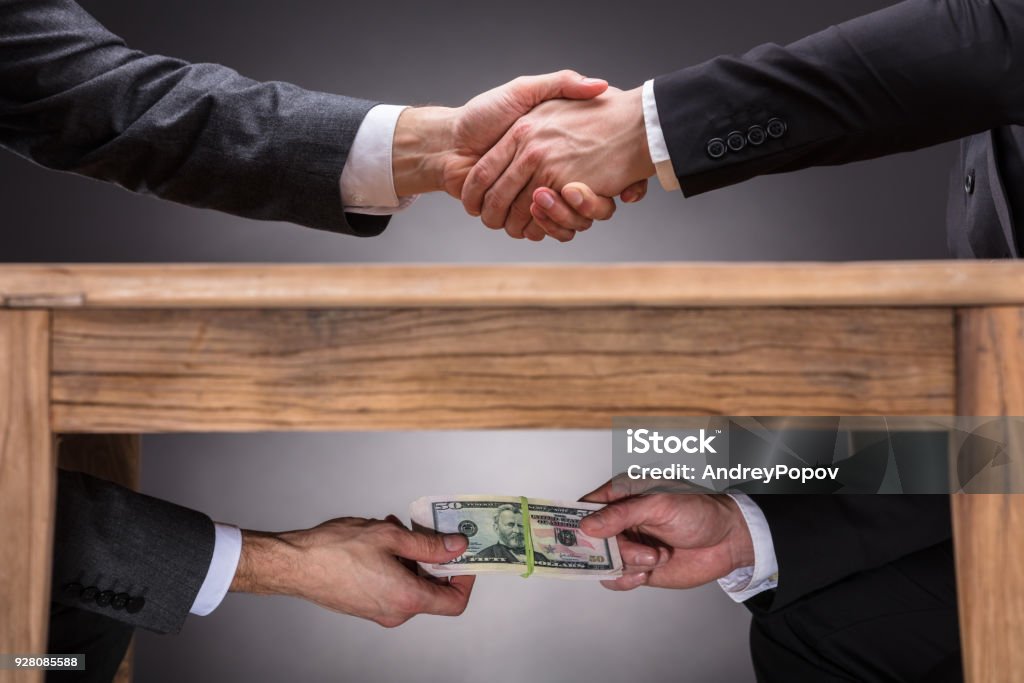 Businesspeople Shaking Hands And Taking Bribe Under Table Close-up Of Two Businesspeople Shaking Hand And Taking Bribe Under Wooden Table On Grey Background Corruption Stock Photo