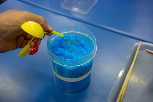 Hands holding spoon of blue chemical, Copper Sulphate Pentahydrate, against blue table for junior scientific lab test and preparation.