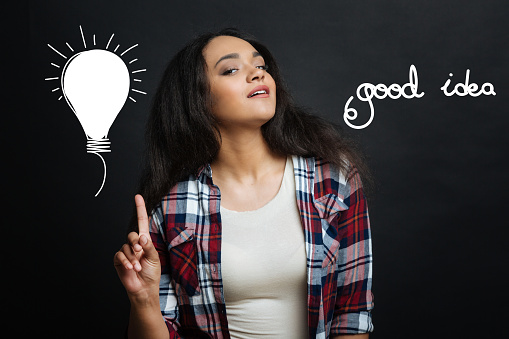 Good idea. Pretty emotional clever woman standing against the gray background and pointing her finger up while thinking about a brilliant idea connected with her amazing project