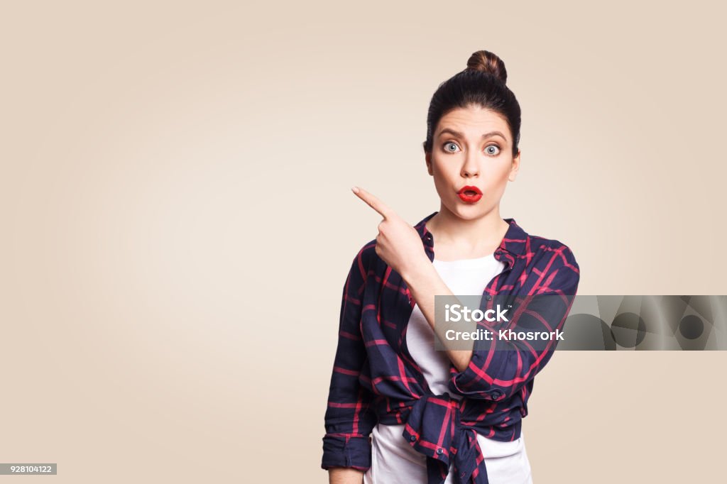 Young surprised girl with casual style and bun hair pointing her finger sideways Young surprised girl with casual style and bun hair pointing her finger sideways, demonstrating something on beige blank wall with copy space for your information or promotional content. Awe Stock Photo
