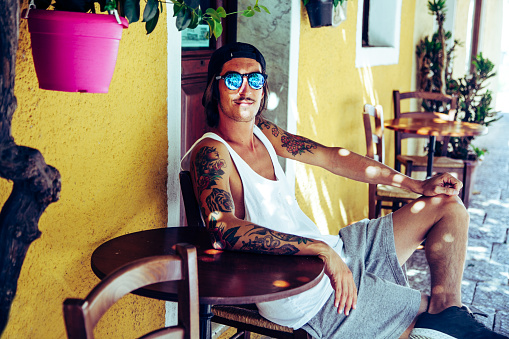 portrait of tattooed boy on holiday in Italy, Sardinia, sitting on a smiling chair in front of a colorful building