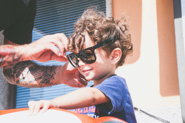 Father with tattoos puts glasses on his 2-year-old, uv sun prevention Father with tattoos puts glasses on his 2-year-old child, prevention of uv rays of the sun occhiali da sole stock pictures, royalty-free photos & images
